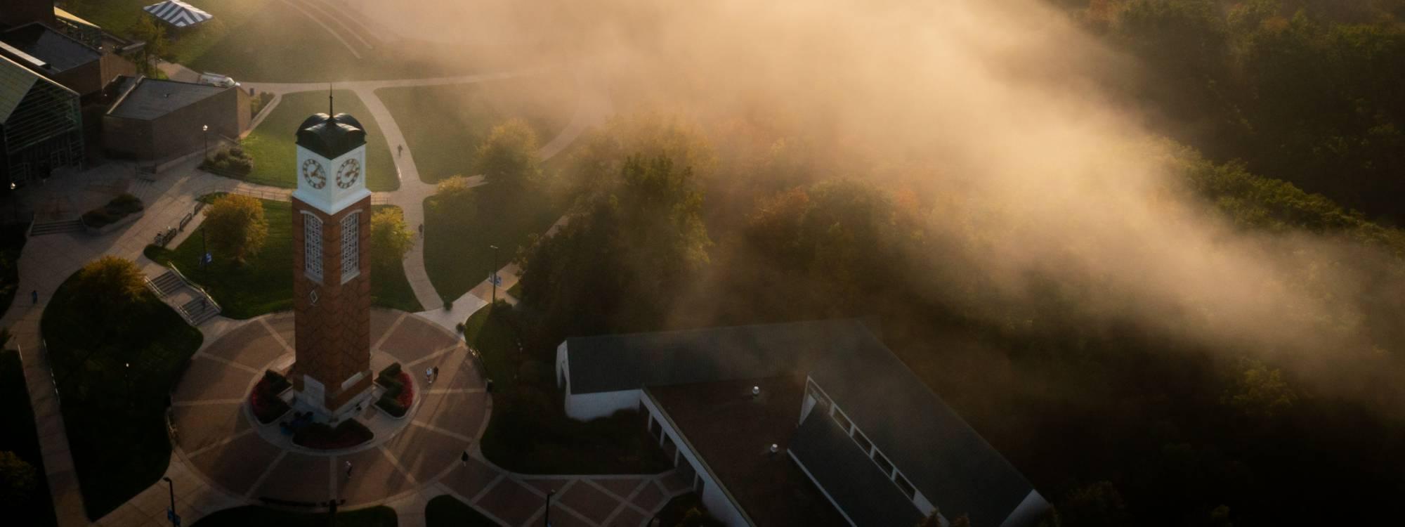 An aerial photo of the Allendale campus shows the clock tower and Cook DeWitt Center as fog rises above the ravines.